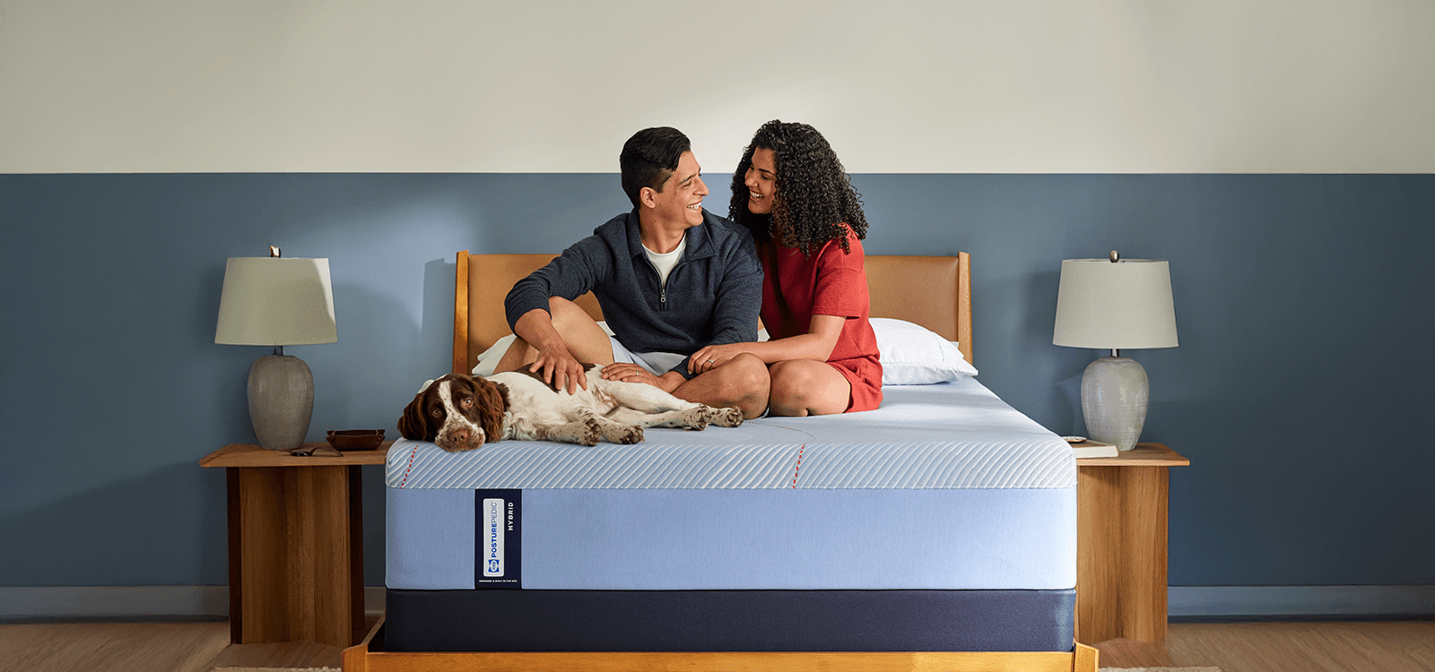 A man and woman sitting on a Posturepedic Hybrid mattress with their dog